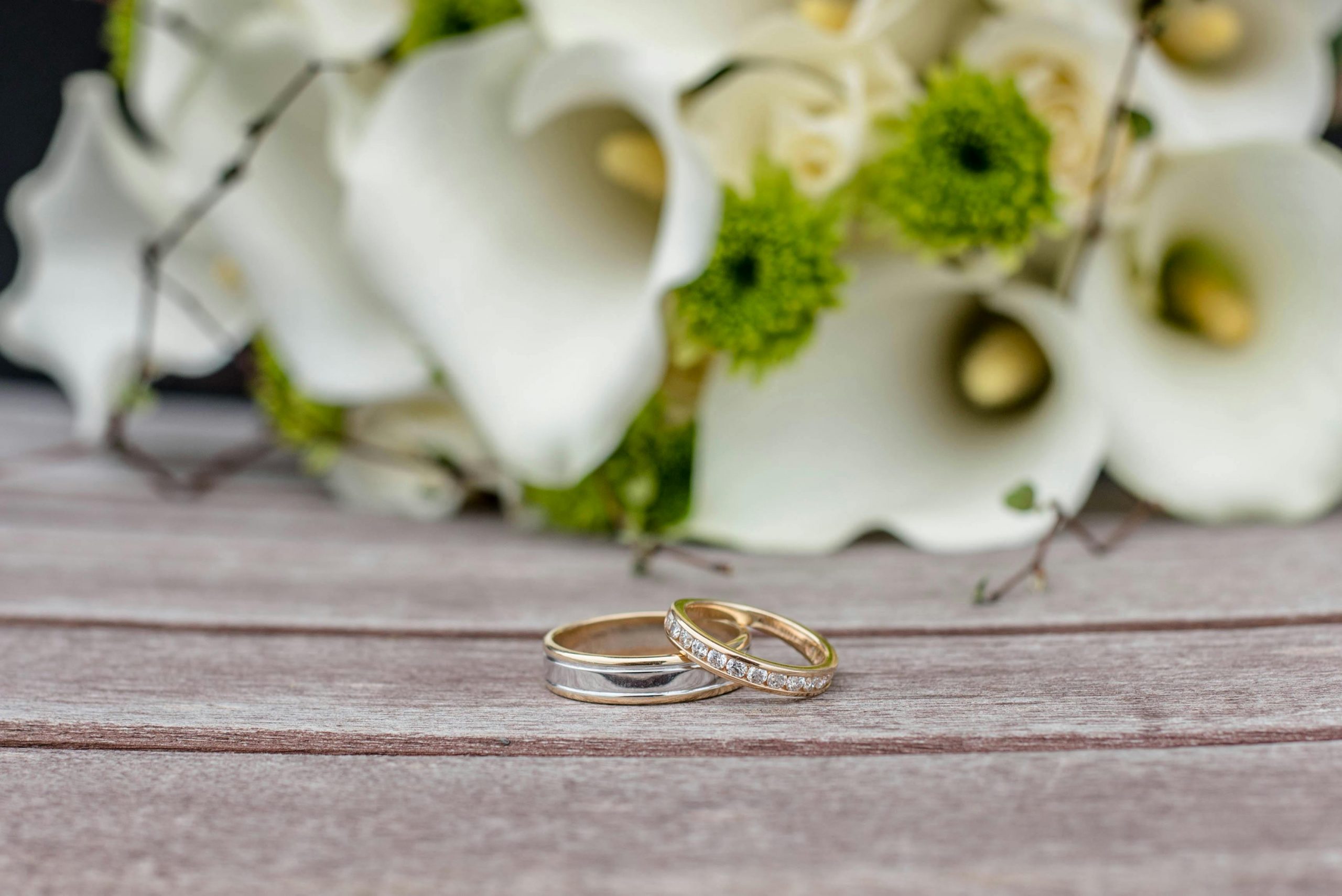 Close-up of wedding rings on wooden surface with white and green floral arrangement.