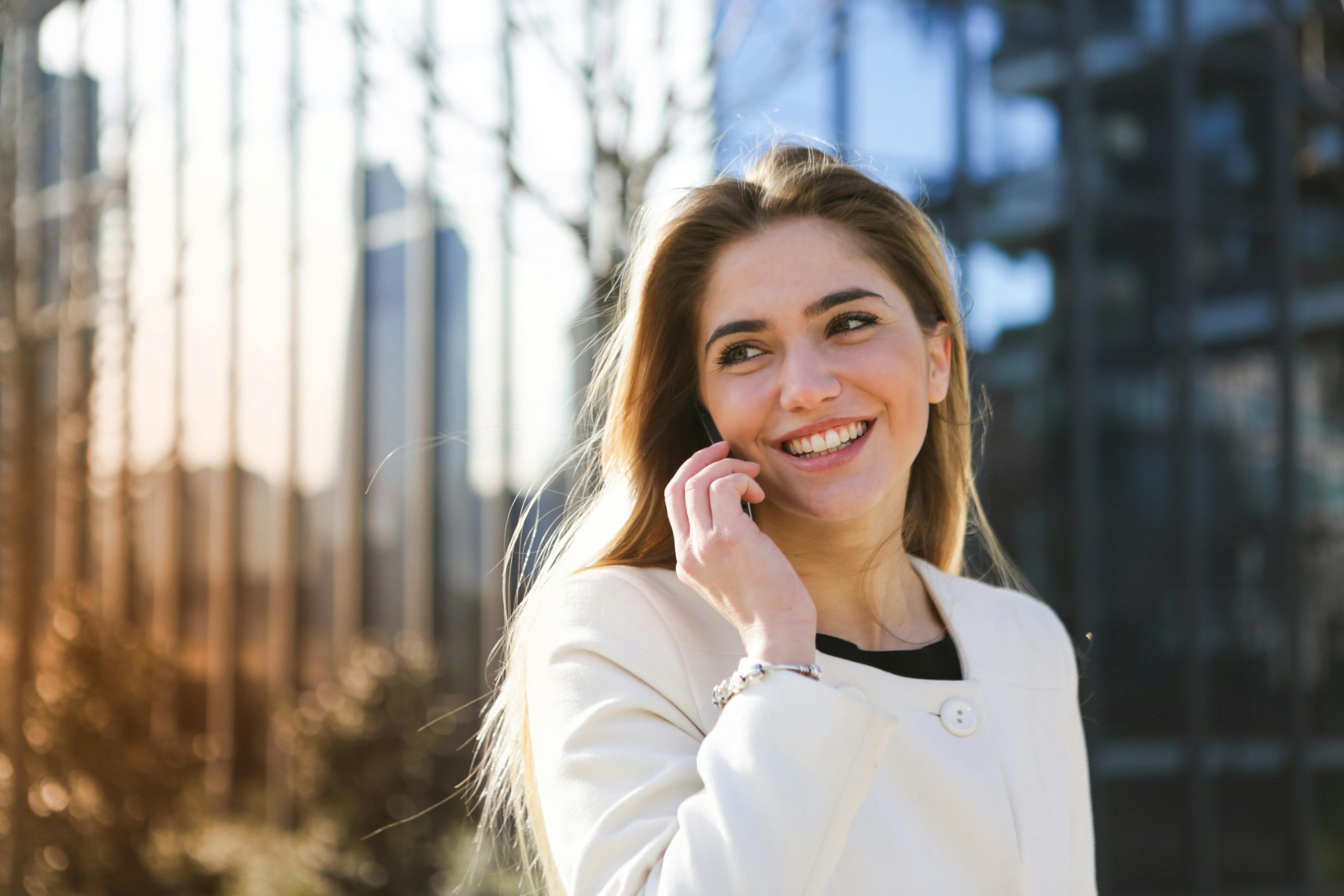 Happy woman in white coat chatting on mobile phone in an urban setting with sunlight.